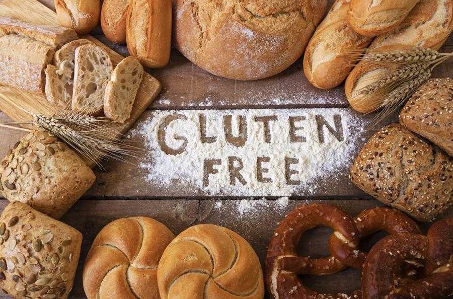 Top view of different types and shapes of gluten free breads on a wood background, forming almost a cirle, and text in the middle written apparently with finger on a white flour that says gluten free